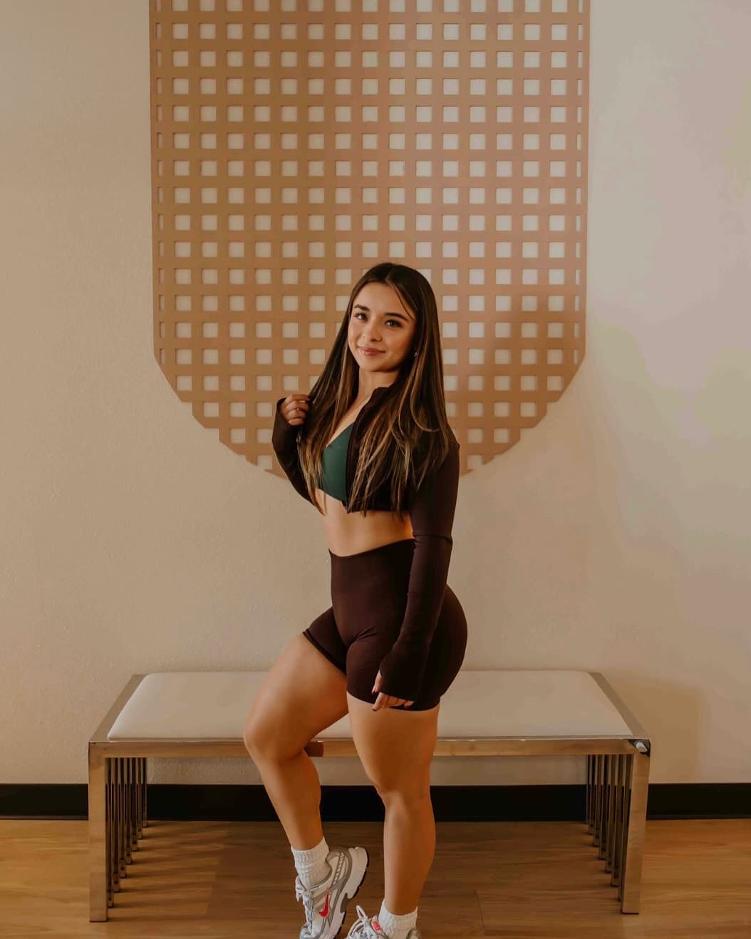 Leslie standing in front of a tan lattice wall wearing a brown workout set